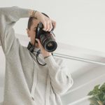 Person in beige sweater holds camera overhead while photographing in minimalist white room with hanging plants, modern chair, and natural window light.