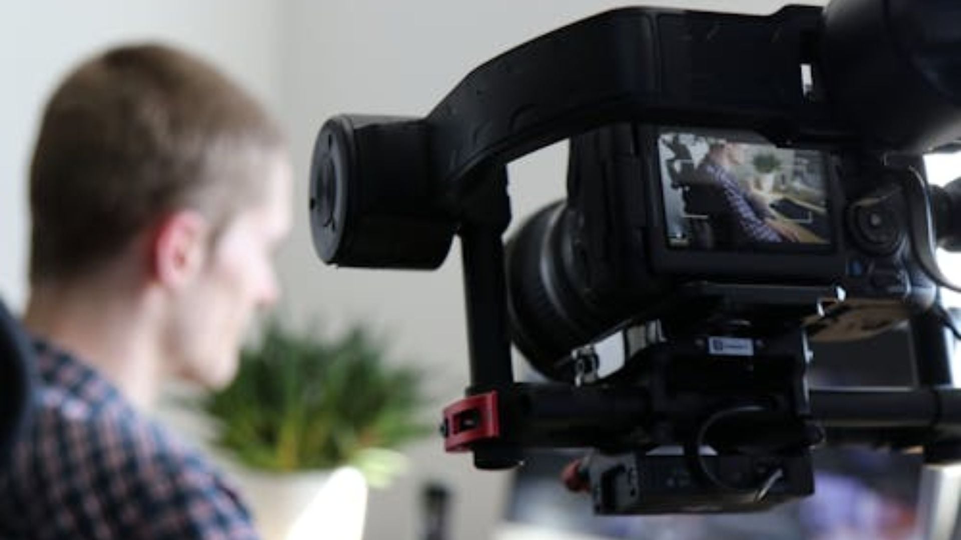 Close-up of professional video camera LCD screen showing subject being recorded during interview, with blurred person and plant visible in background studio.