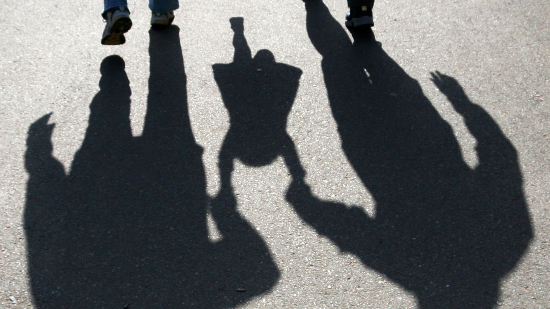 Multiple overlapping shadows on pavement showing adults and small child holding hands together with person's feet visible at top of frame.