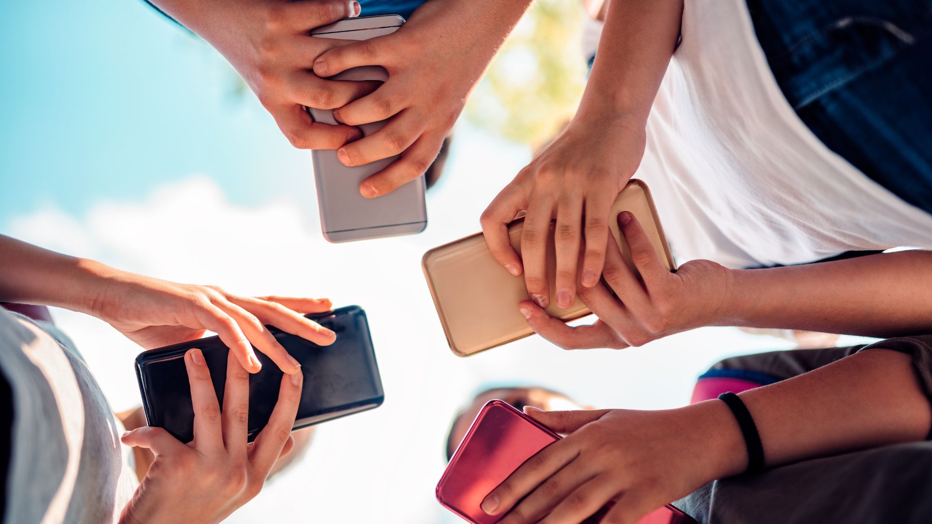 Overhead view of diverse hands holding multiple smartphones in various colors including black, gold, and red gathered in circle outdoors.