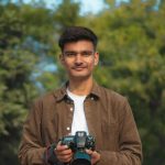 Young man in brown shirt smiles while holding professional DSLR camera with blue accent standing in green outdoor park setting.