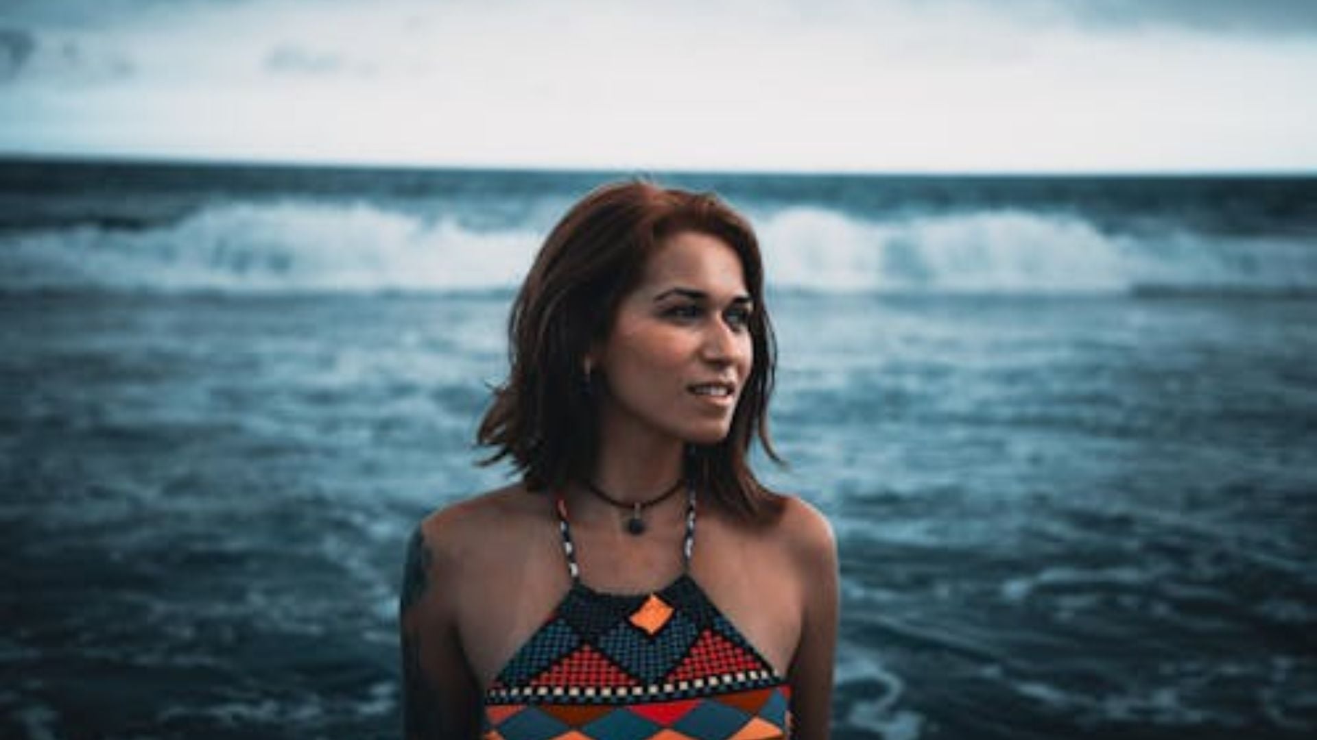 Woman with shoulder-length brown hair wearing colorful geometric patterned halter top with black choker necklace smiling at beach with ocean waves in background.