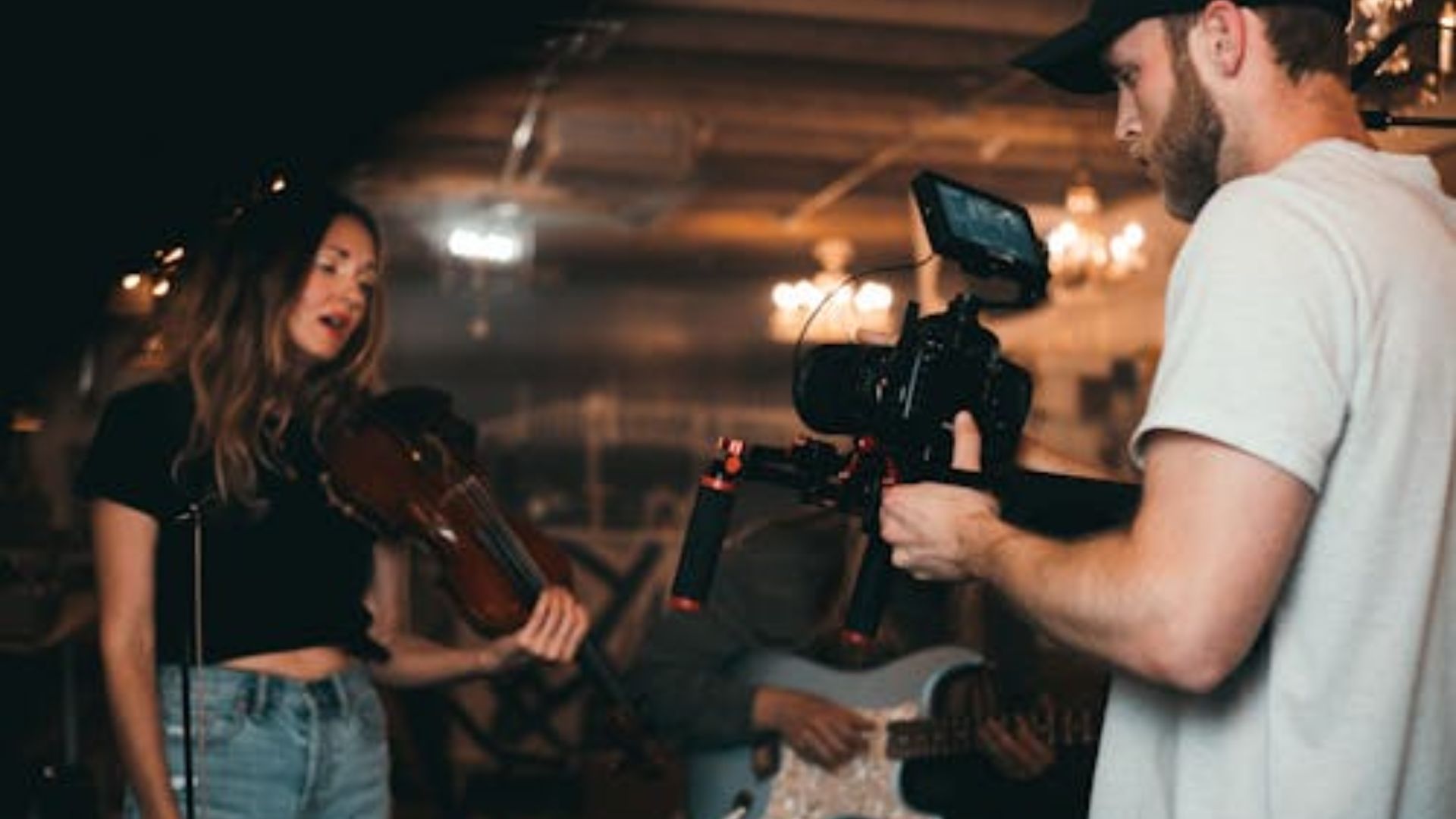 Videographer in white shirt uses handheld gimbal stabilizer to film female musician performing with guitar outdoors at night with cafe lights background.