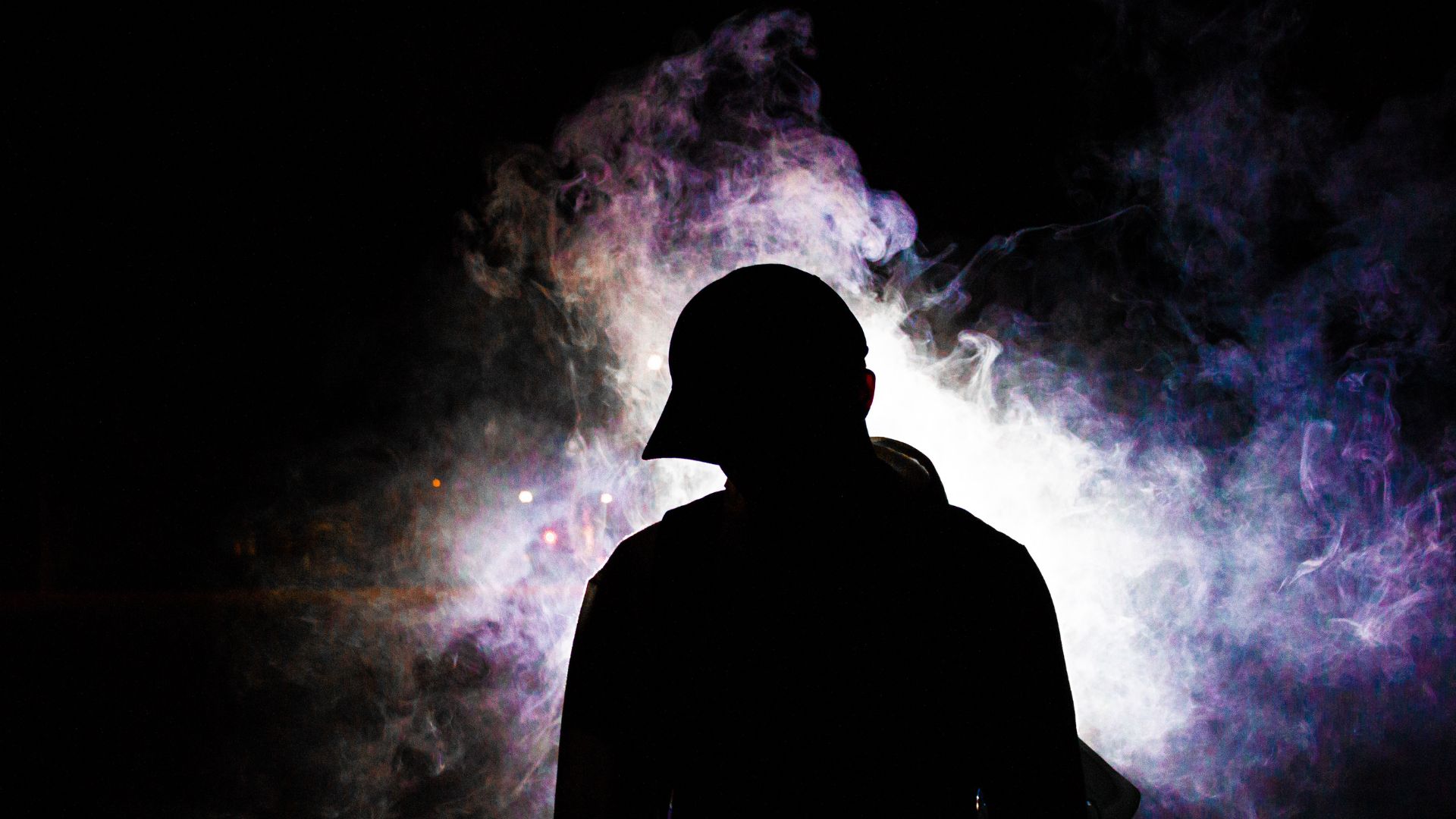 Dark figure silhouette in profile against black background surrounded by swirling purple, blue, and white colored smoke or vapor clouds.