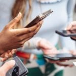 Close-up of woman's hands holding rose gold smartphone with other people using their devices blurred in background at social gathering.