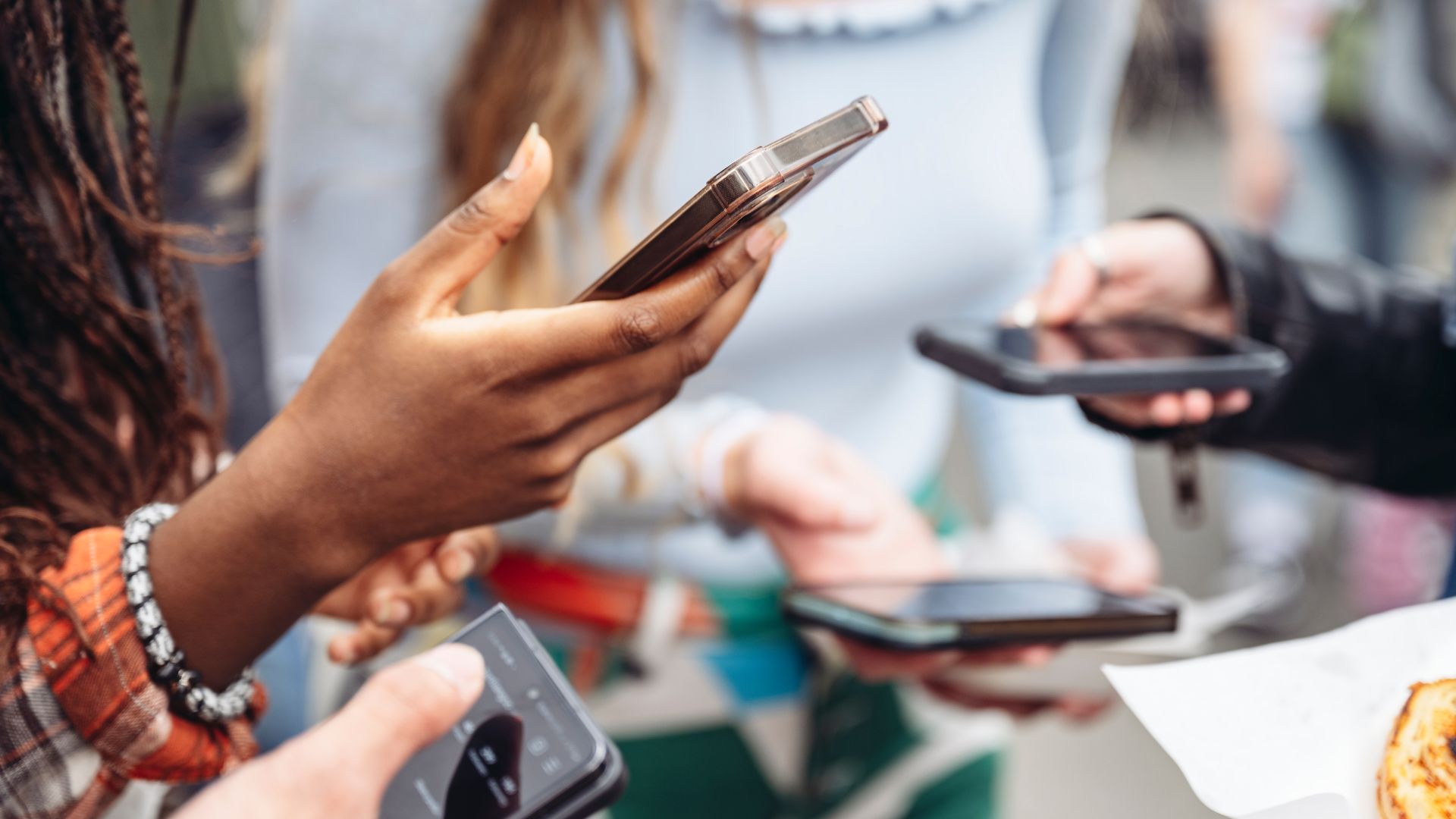 Close-up of woman's hands holding rose gold smartphone with other people using their devices blurred in background at social gathering.