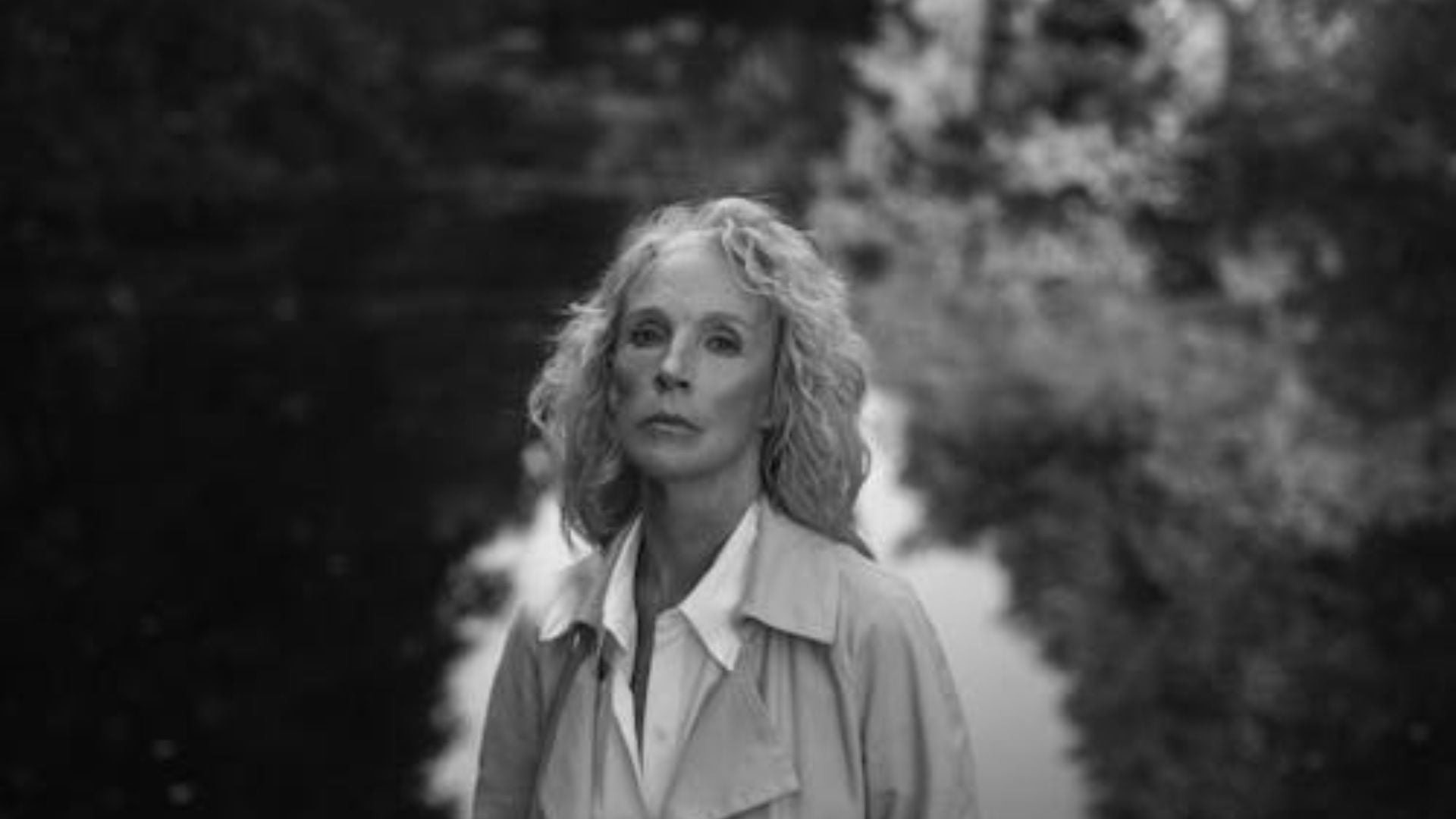 Artistic black and white portrait of woman with wavy blonde hair wearing light collared jacket looking upward outdoors with soft bokeh natural background.