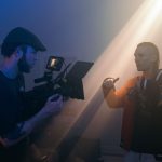 Cameraman with gimbal rig films woman in orange vest against dramatic blue and gold studio lighting creating atmospheric indoor production scene with haze.