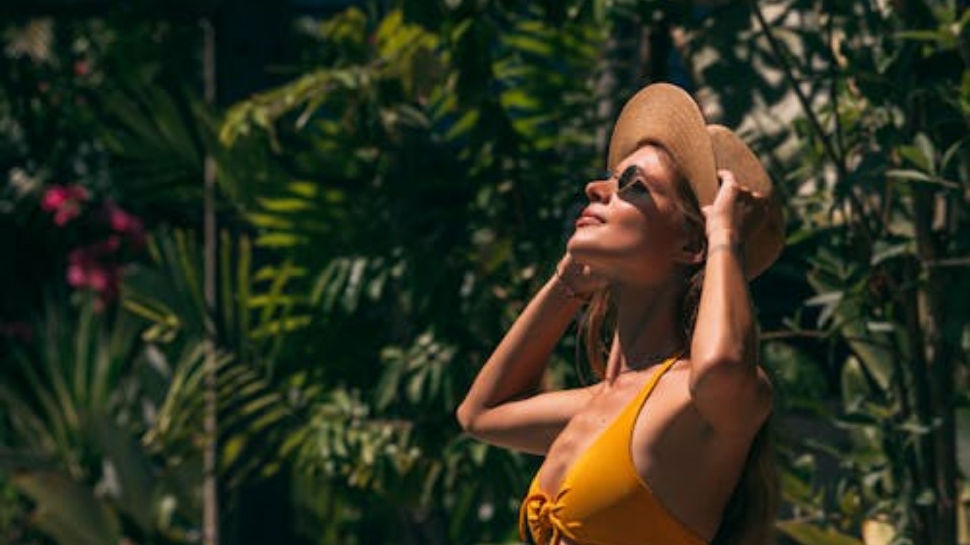 Woman wearing yellow swimsuit and wide-brim straw hat with sunglasses looking up at sky outdoors with lush green tropical garden plants in background.