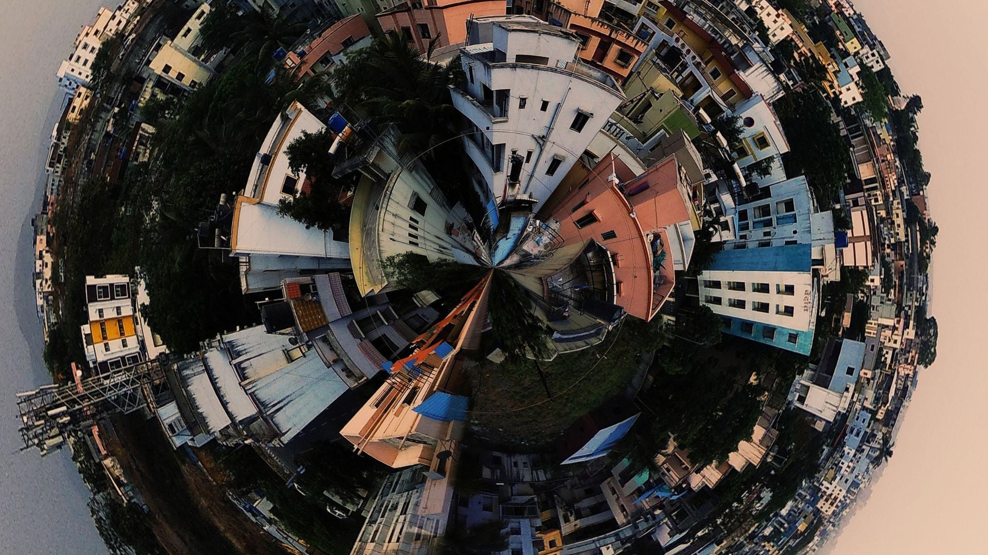 Spherical tiny planet 360 panorama showing dense urban residential area with colorful buildings, rooftops, narrow streets, and trees warping around central point in creative fisheye distortion effect.