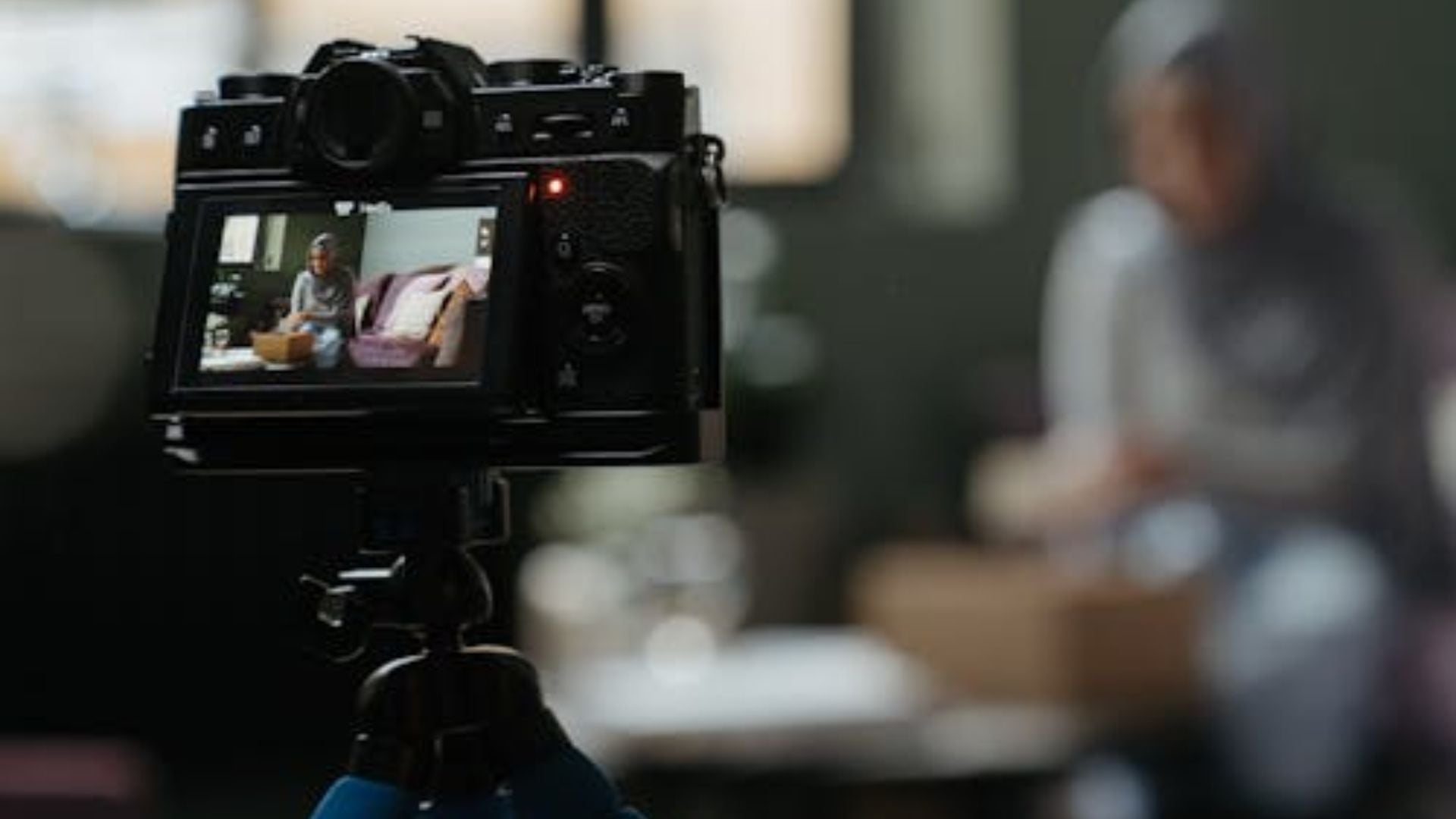 Black digital camera LCD screen displays woman sitting on pink couch during video production, with blurred production crew visible in foreground.