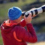 Photographer wearing blue knit hat and red jacket using large white telephoto lens on camera outdoors in natural mountain landscape setting.