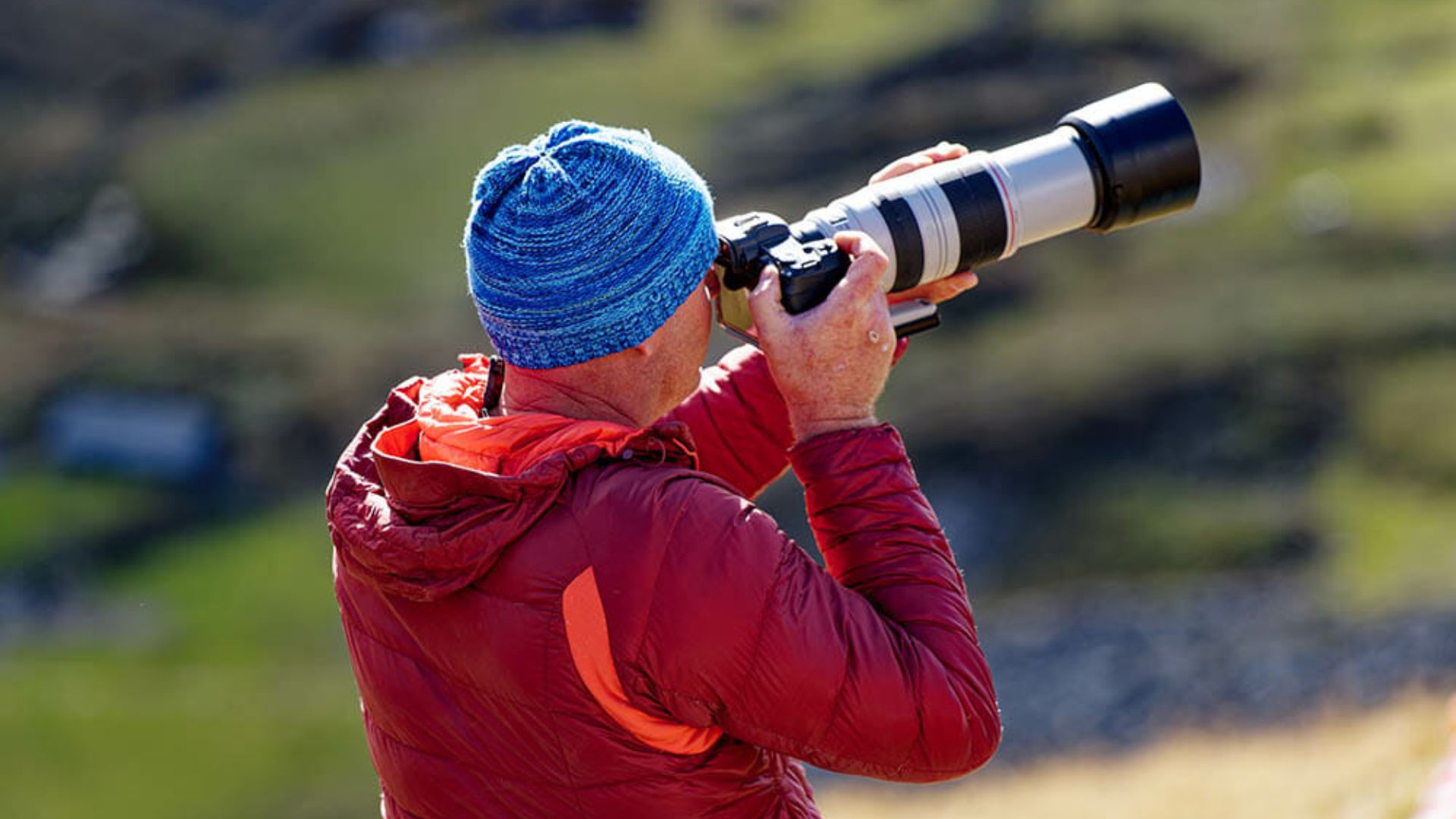 Photographer wearing blue knit hat and red jacket using large white telephoto lens on camera outdoors in natural mountain landscape setting.