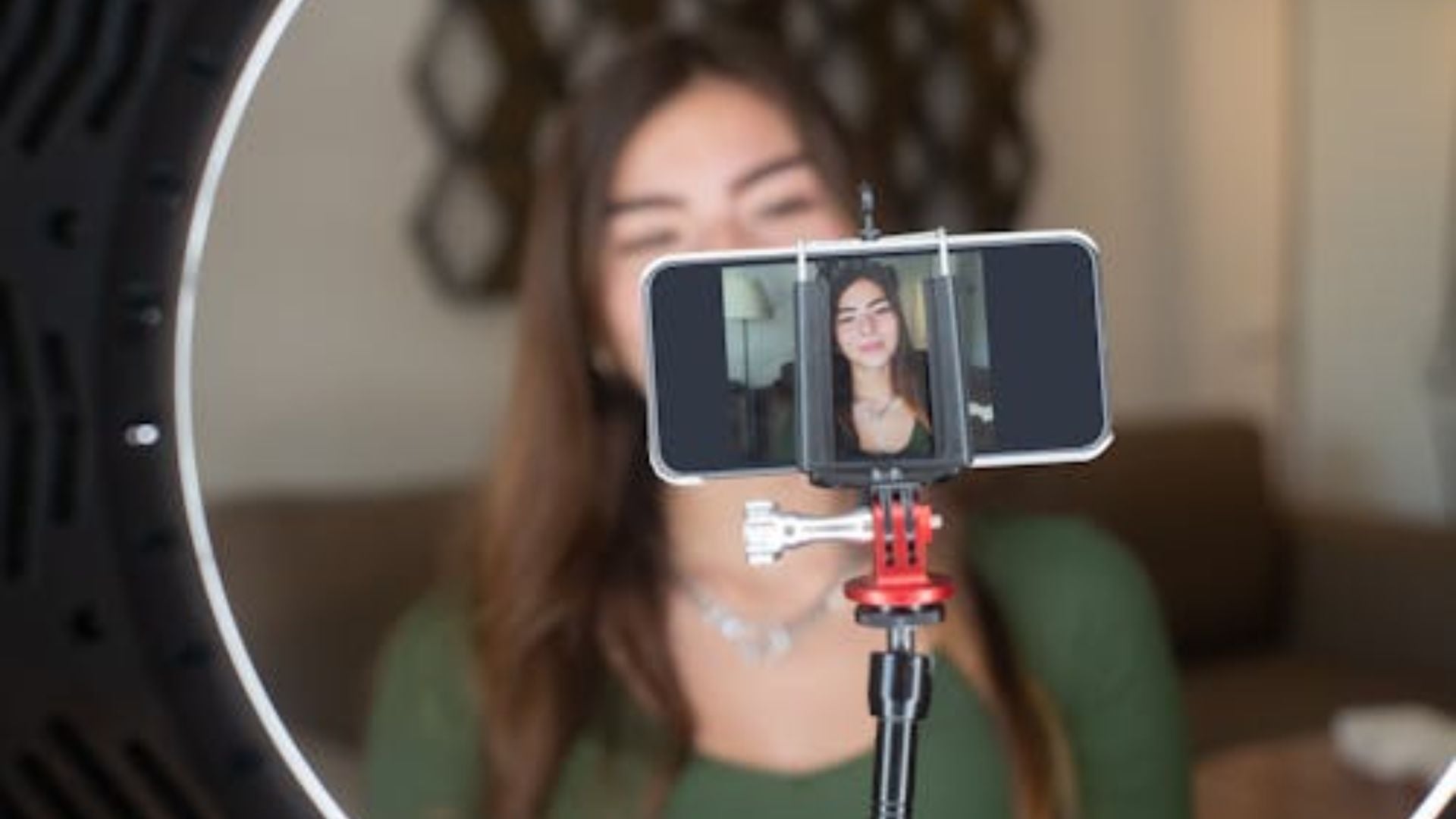 Smartphone mounted on small tripod with red adapter filming woman in green shirt, ring light visible at edge creating professional home recording setup.