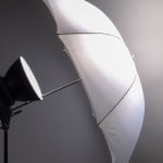 Black strobe light with white translucent umbrella modifier mounted on stand creates soft diffused lighting for professional photography against neutral backdrop.