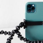 Turquoise iPhone Pro secured in black flexible tripod with bendable legs and adjustable grip clamp on white surface for versatile positioning.
