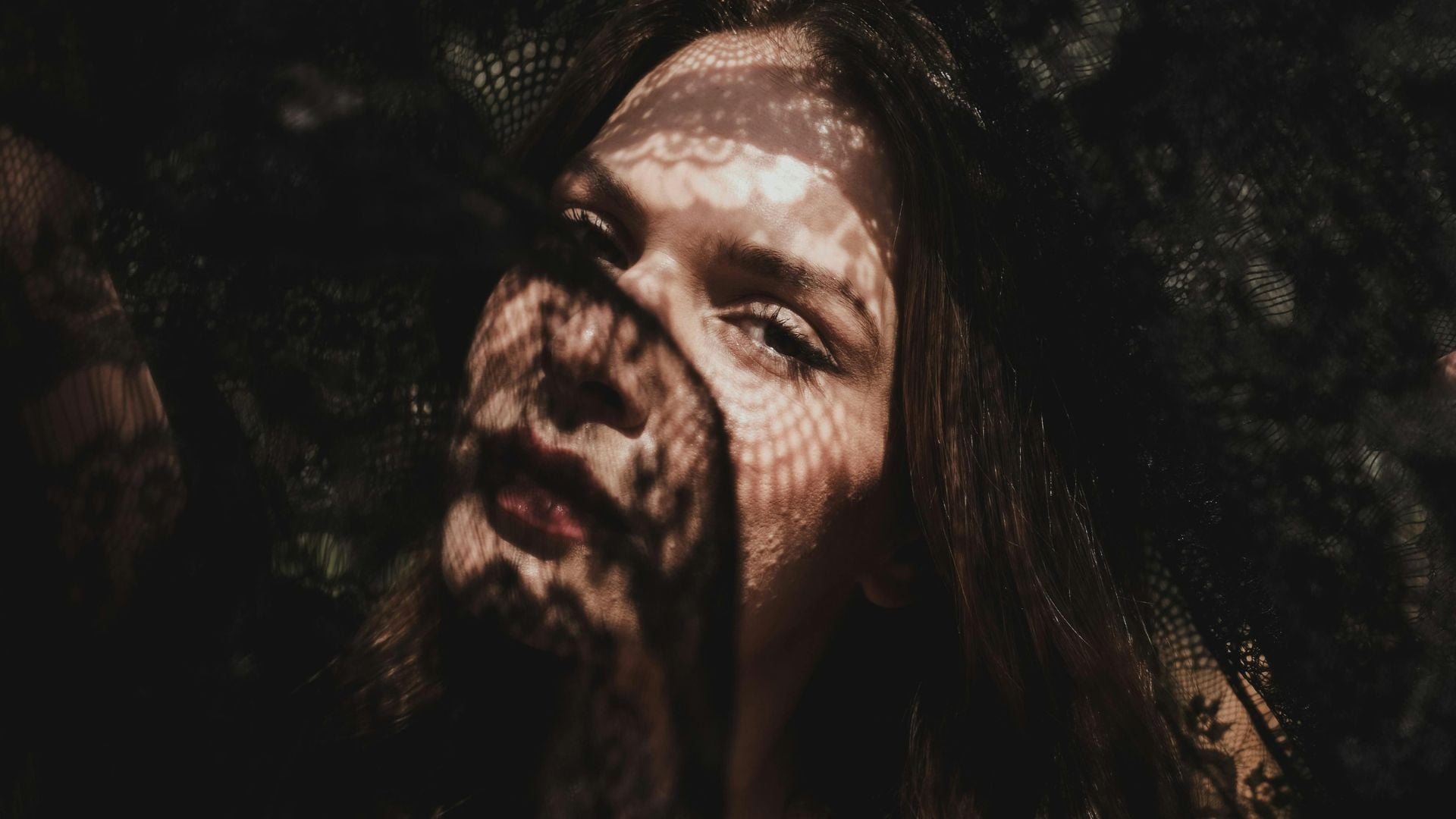 Close-up portrait of woman looking upward with intricate lace or mesh shadow pattern creating artistic dappled light across her face.