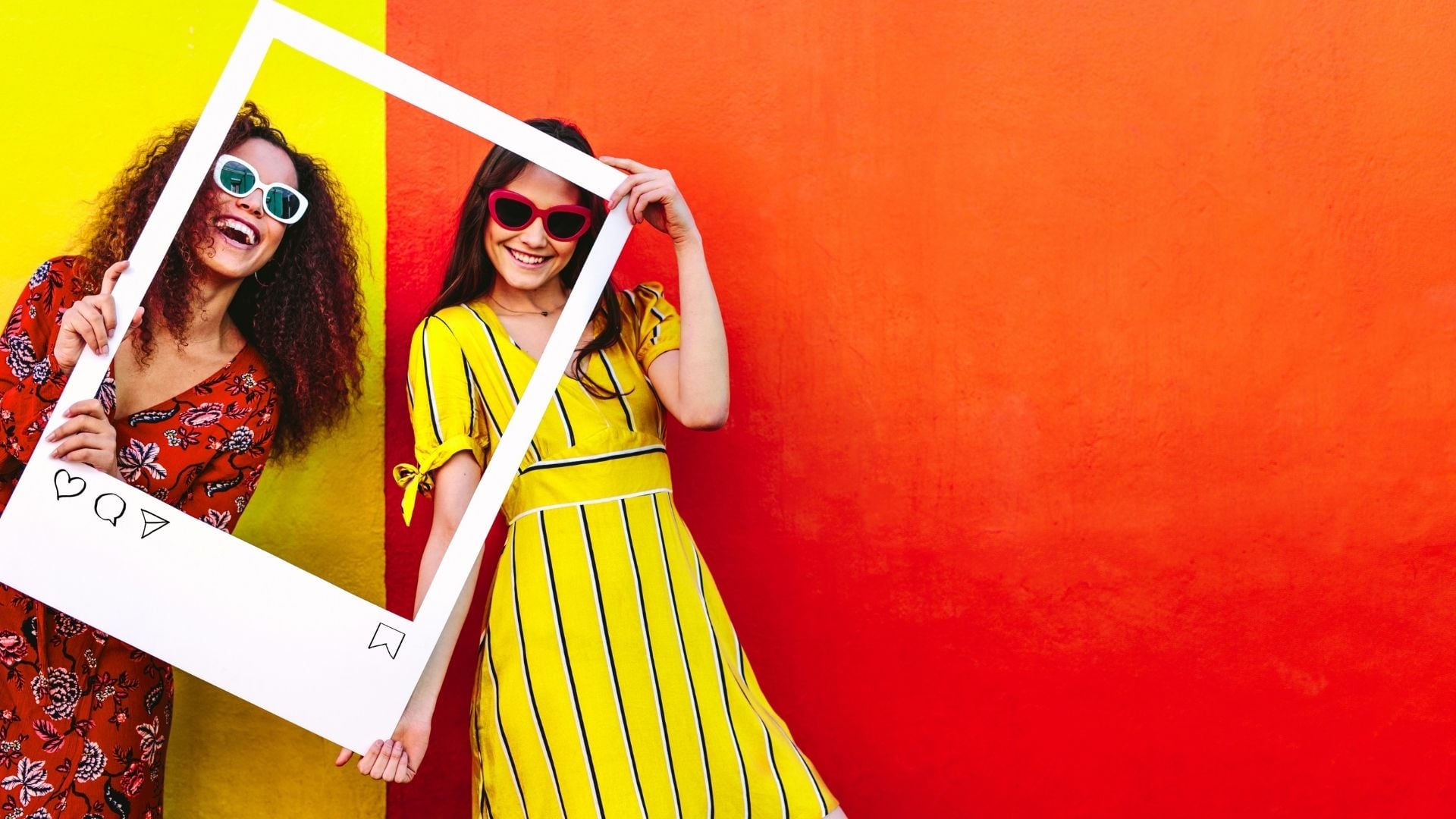 Two smiling women wearing colorful dresses and sunglasses holding large white Instagram-style frame prop against vibrant yellow and red split background for social media content creation.