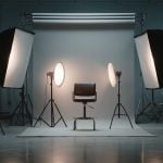 Complete photography studio featuring white backdrop, chair subject placement, four softbox lights on tripods, two circular LED lights, overhead backdrop system in industrial space with blue walls.