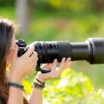 Woman with long brown hair wearing beaded bracelets using black telephoto lens camera in bright outdoor garden setting with green foliage background, demonstrating handheld telephoto photography technique.