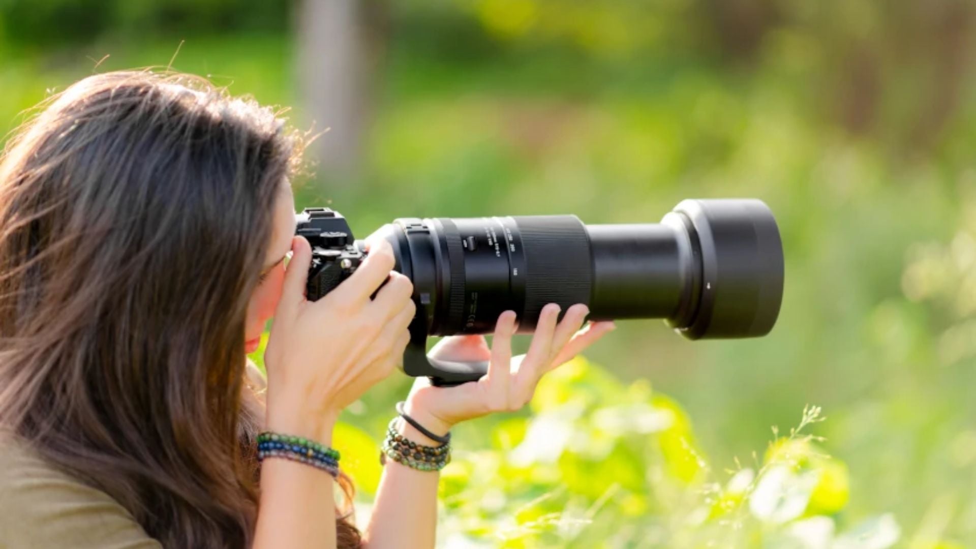 Woman with long brown hair wearing beaded bracelets using black telephoto lens camera in bright outdoor garden setting with green foliage background, demonstrating handheld telephoto photography technique.