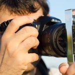 Photographer holding Nikon DSLR camera up to eye with glass prism held in front of lens, creating creative light refraction effects outdoors.