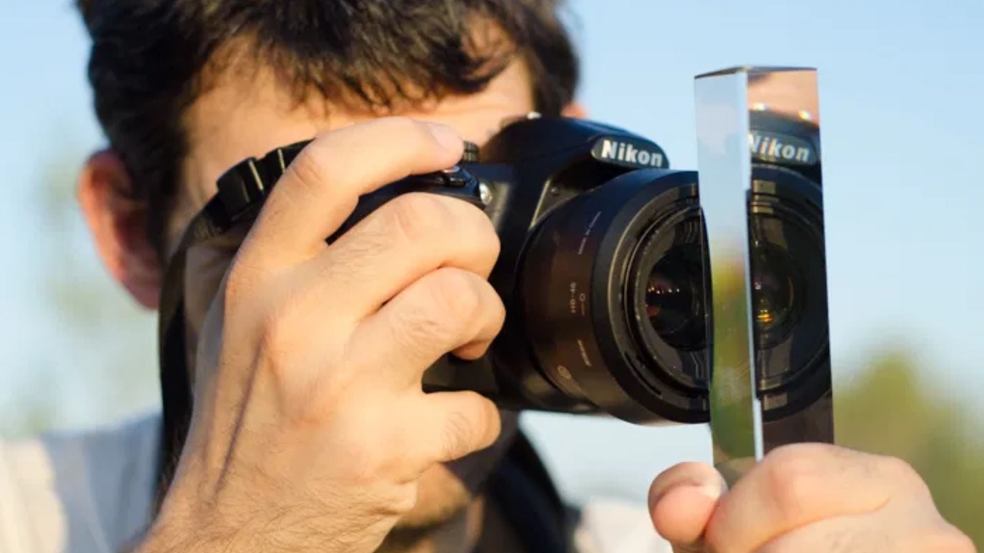 Photographer holding Nikon DSLR camera up to eye with glass prism held in front of lens, creating creative light refraction effects outdoors.