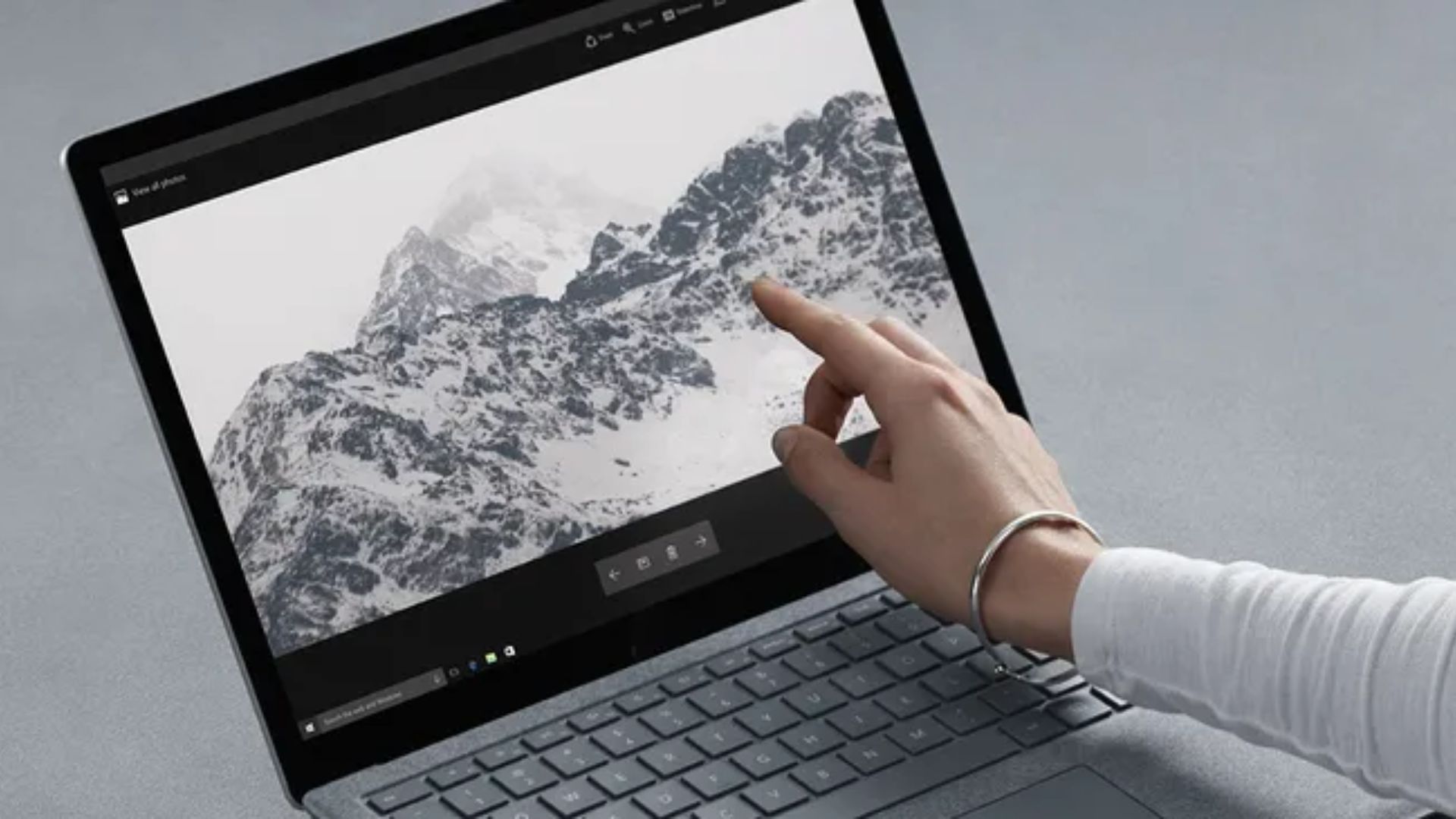 Hand with silver bracelet touching touchscreen laptop displaying black and white snowy mountain landscape photo, demonstrating interactive editing interface with visible keyboard below screen.