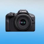 Black Canon EOS R100 mirrorless camera with RF-S 18-45mm kit lens mounted showing front view with textured grip on blue gradient background.