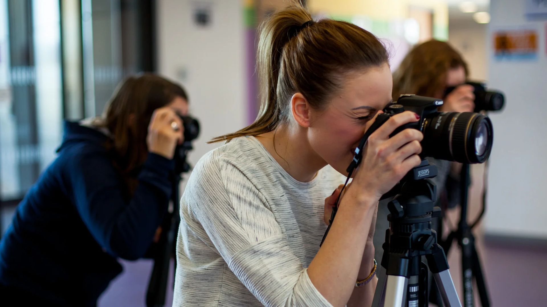 Woman with ponytail in beige sweater looking through Canon DSLR camera viewfinder on tripod in photography class, other students visible shooting in blurred background studio setting.