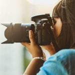 Woman in blue top with bracelet holds DSLR camera with telephoto lens up to face, photographing outdoors in soft natural light with bokeh background.