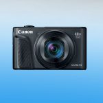 Black Canon PowerShot SX740 HS compact camera front view showing 40x optical zoom lens, textured grip, brand logo, and model name on light blue background.