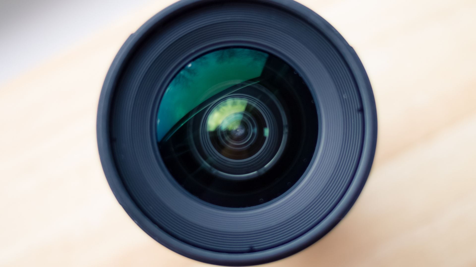 Macro view of fisheye camera lens showing curved front glass element with green and yellow reflections, circular ribbed lens hood, professional photography equipment on neutral background.