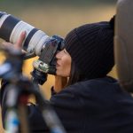Female photographer wearing black winter hat and hoodie using professional white telephoto lens mounted on monopod with blurred autumn background, capturing wildlife or sports photography action.