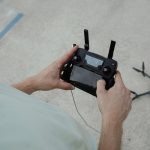 Person's hands holding black drone remote controller with LCD display screen and folding antennas over concrete pavement, drone shadow visible on ground, demonstrating outdoor aerial photography operation.