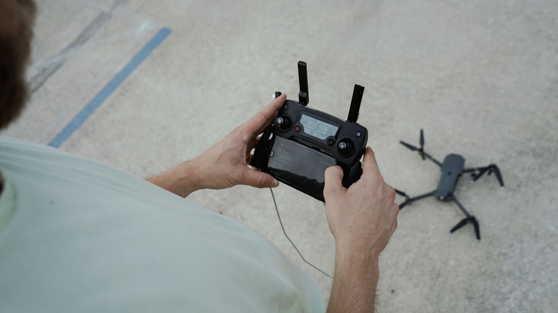 Person's hands holding black drone remote controller with LCD display screen and folding antennas over concrete pavement, drone shadow visible on ground, demonstrating outdoor aerial photography operation.