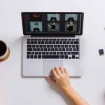 Overhead view of person's hands on MacBook laptop displaying photo gallery grid with six portrait images, wooden desk with striped coffee mug, smartphone, colorful paperclips, SD card, DSLR camera, and yellow-spine notebook.