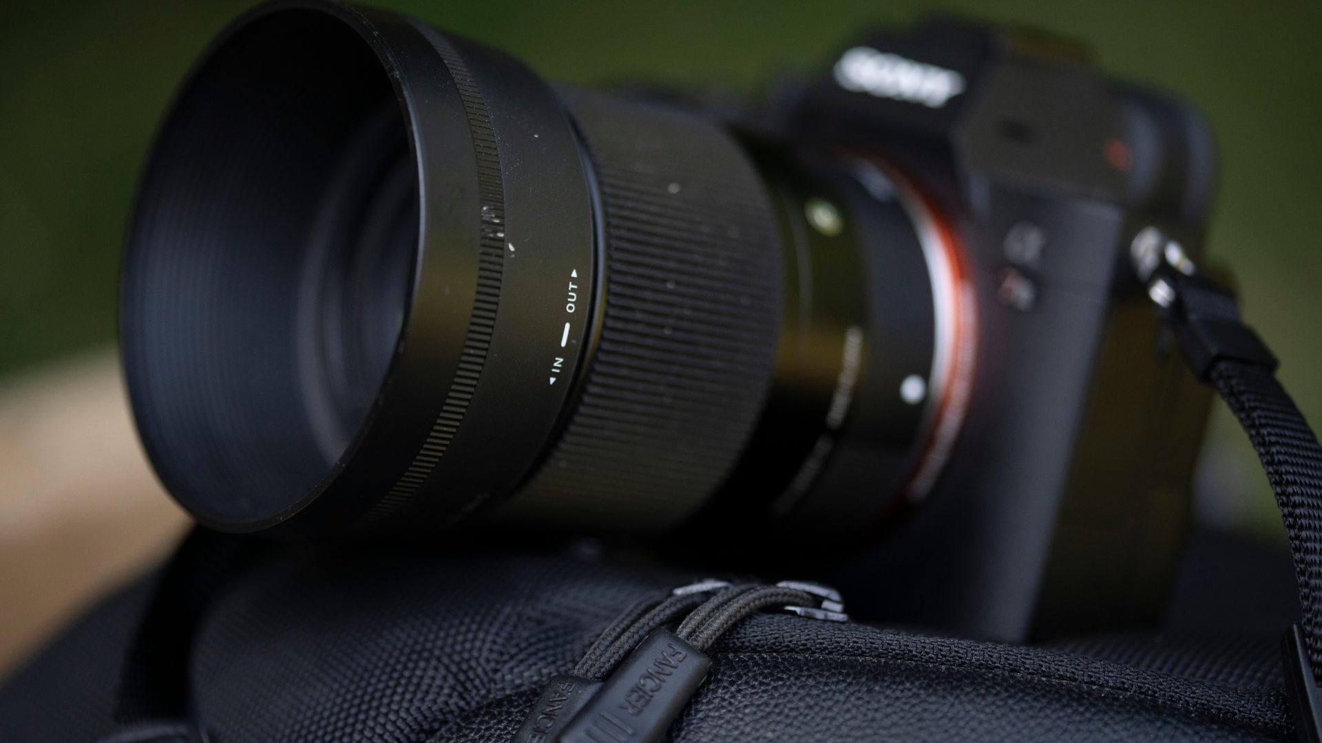 Black telephoto lens with visible IN-OUT markings on focus ring attached to blurred camera body resting on textured black camera strap outdoors.