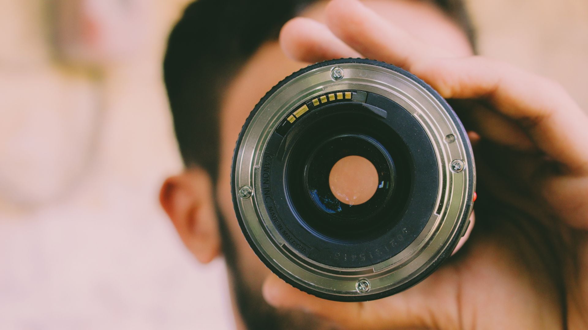 Hand holding camera lens showing rear mount with silver metal ring, electronic contact pins, glass elements visible, central aperture opening, blurred photographer background demonstrating lens attachment.