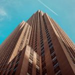 Extreme low-angle wide shot of tall brown concrete skyscraper tower converging toward blue sky with white contrail, dramatic vertical perspective emphasizing height and modern urban architecture.