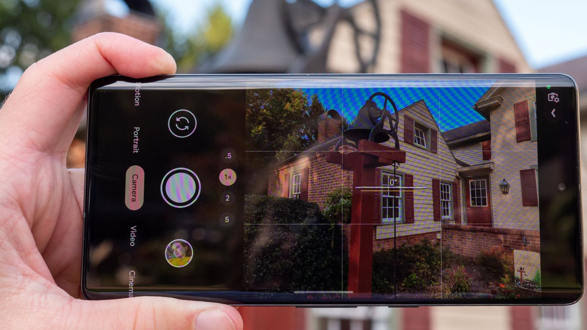 Hand holding phone horizontally showing GCam interface with portrait mode, camera controls, zoom options, and house with colorful lighting effects displayed on screen.