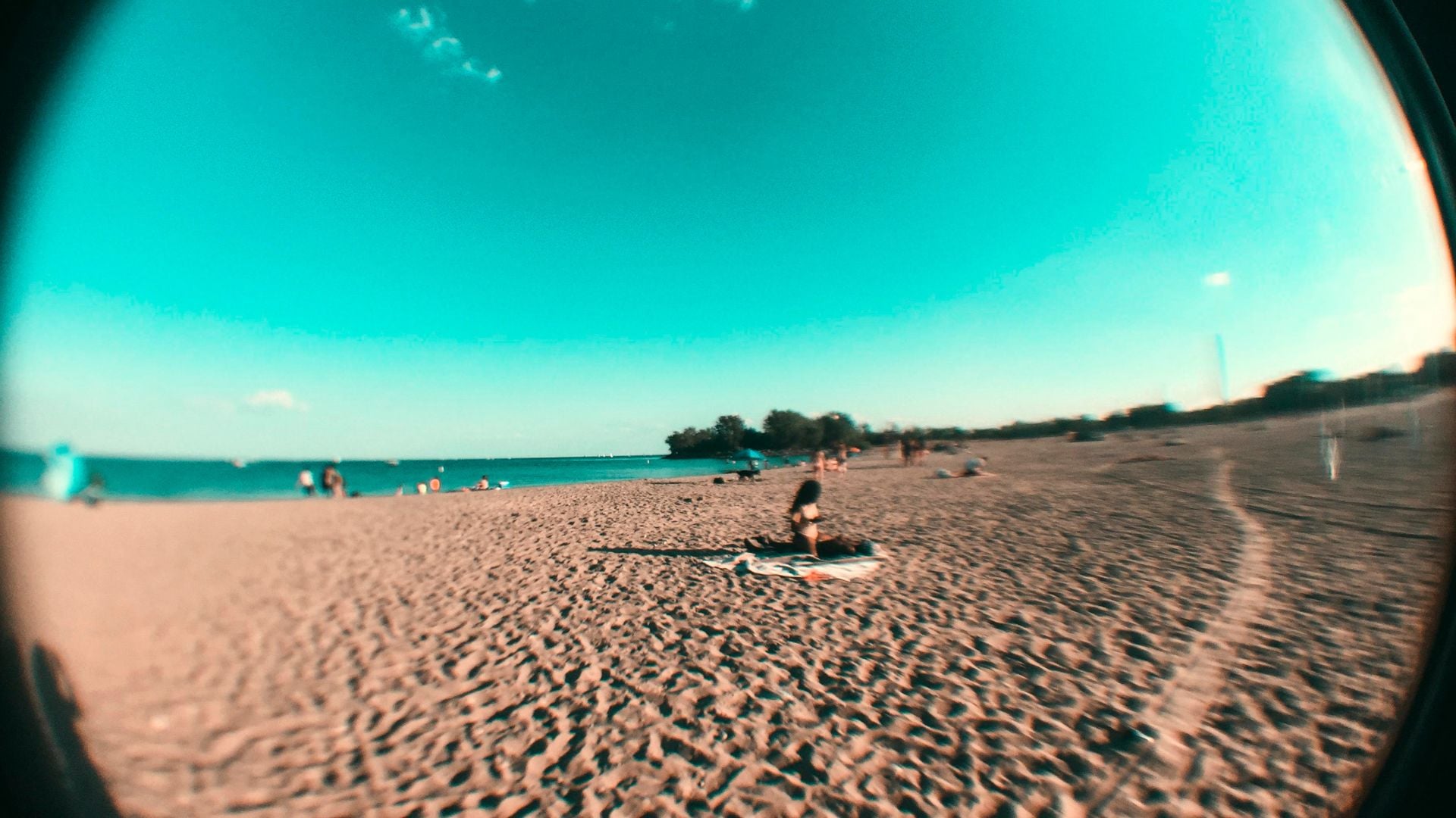 Fisheye lens beach photograph showing curved turquoise sky, sandy shore, person relaxing on towel, distant beachgoers, turquoise water, coastal trees creating ultra-wide distorted circular perspective.