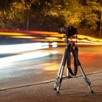 Camera on tripod capturing vehicle light trails at night. Red and yellow streaks show long exposure technique with autumn trees and roadway creating colorful movement effect.
