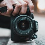 Hand wearing silver ring holds black Canon DSLR camera with EF-S 18-55mm kit zoom lens on wet pavement with warm bokeh background.