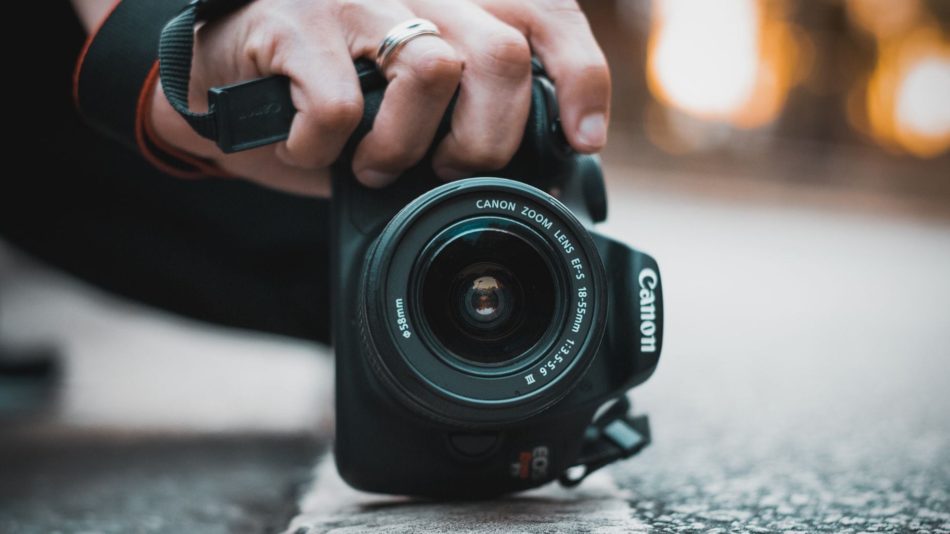 Hand wearing silver ring holds black Canon DSLR camera with EF-S 18-55mm kit zoom lens on wet pavement with warm bokeh background.