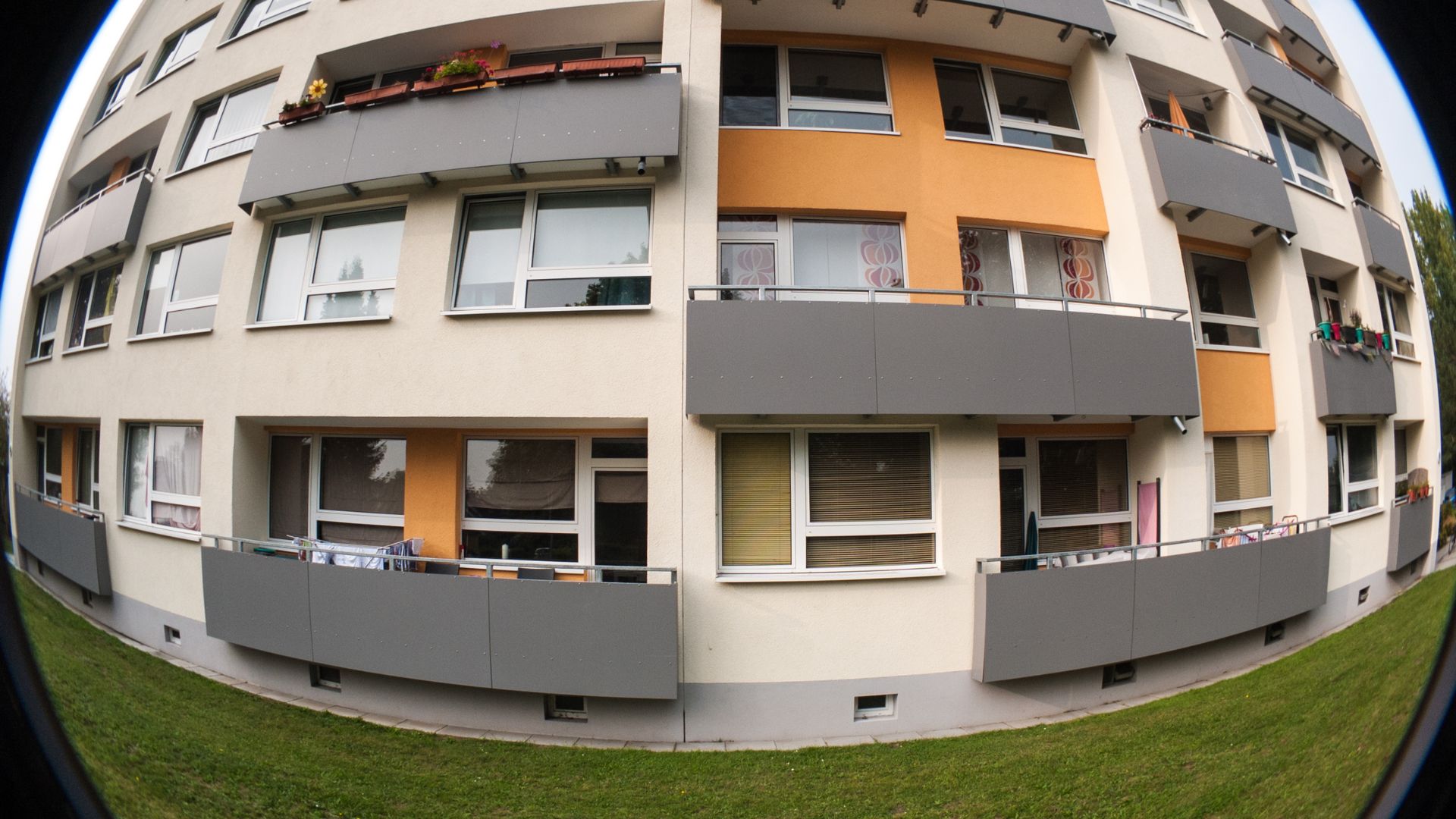 Fisheye photograph of curved multi-story residential building with gray balconies, orange accent panels, white windows, green lawn foreground, creating dramatic architectural barrel distortion perspective.