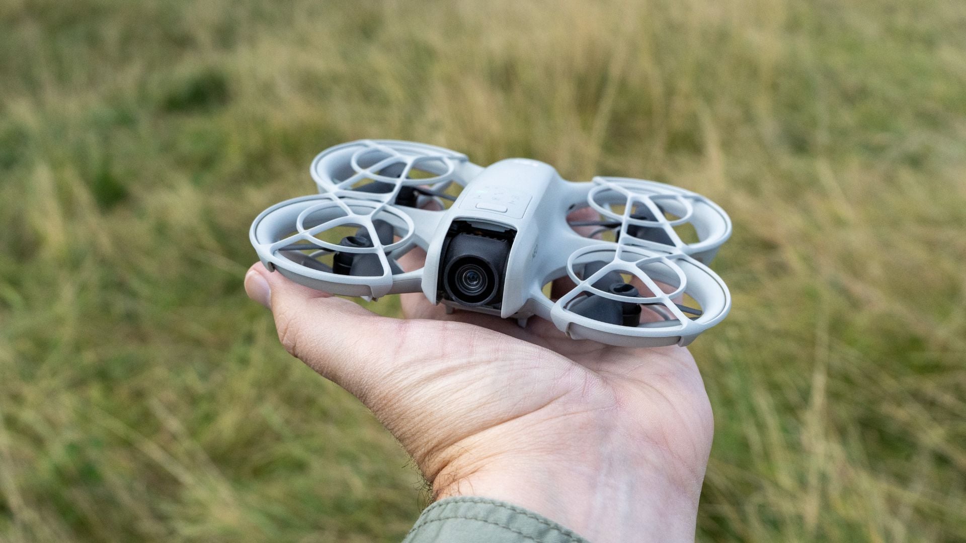 Hand holding white compact FPV racing drone with circular protective propeller guards and front-facing camera outdoors in field with dry grass, showcasing lightweight aerial photography and racing drone design.