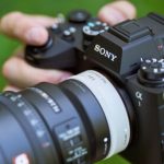 Hands holding Sony mirrorless camera body with attached macro lens against blurred green natural background, showing top controls, viewfinder, and camera strap for outdoor macro photography shooting.