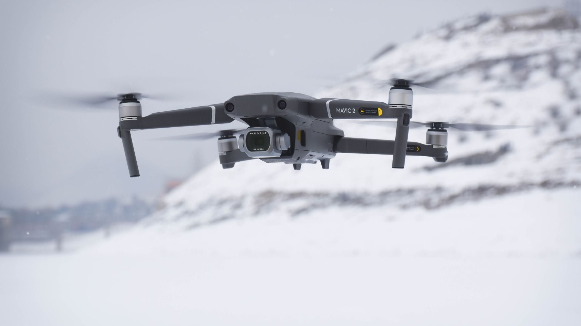 Gray DJI Mavic 2 drone with Hasselblad camera hovering in cold winter environment with blurred snow-covered mountains visible in distant background.