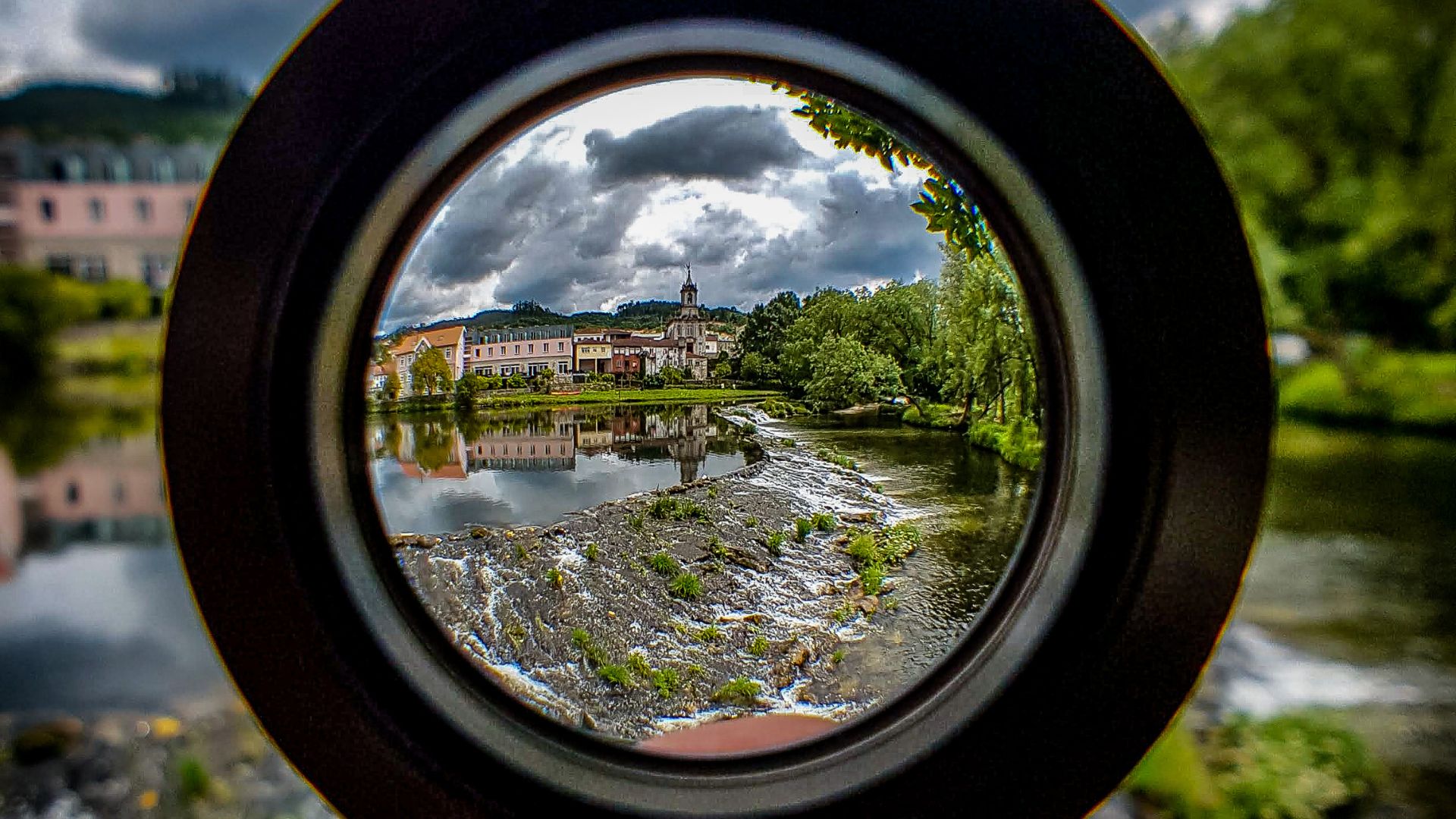 Historic European riverside town viewed through circular fisheye lens showing colorful buildings, church spire, river reflection, dramatic cloudy sky, green trees creating artistic framed landscape composition.